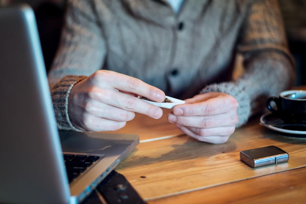 man rolling joint on his desk next to computer and cup of coffee